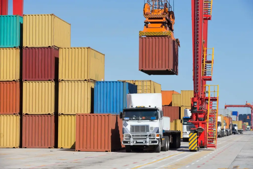 Container crane lifting a container above stacked containers, with a truck in front